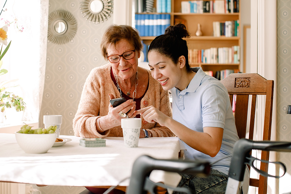Caregiver and elderly woman looking at a mobile phone while sitting at a dining table